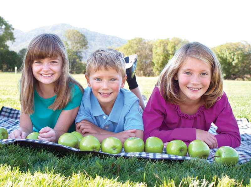 Two young girls and a boy laying outside on a blanket with apples.